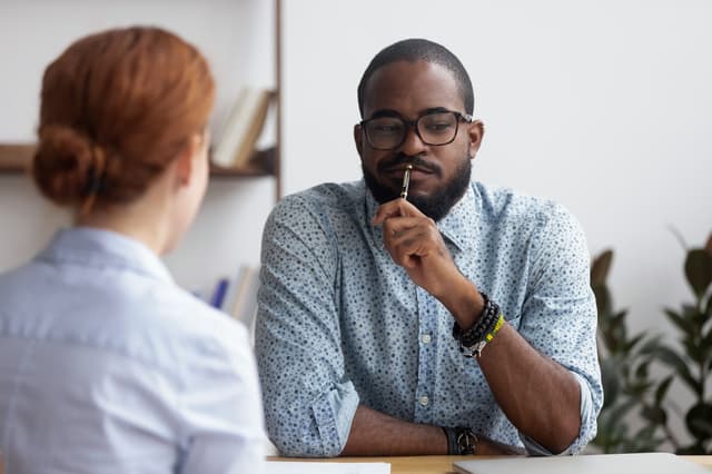 Interviewer in glasses holding a pen while listening to a candidate at a desk