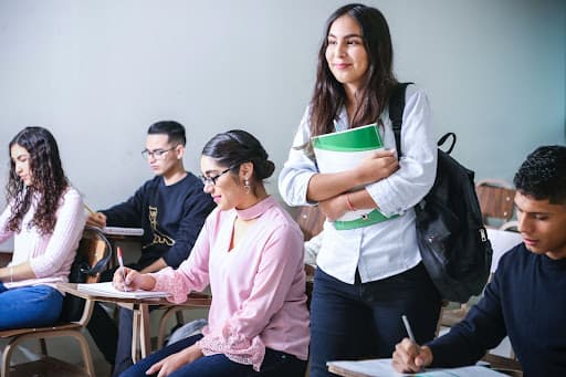 Student holding notebooks and a backpack standing among classmates writing at desks