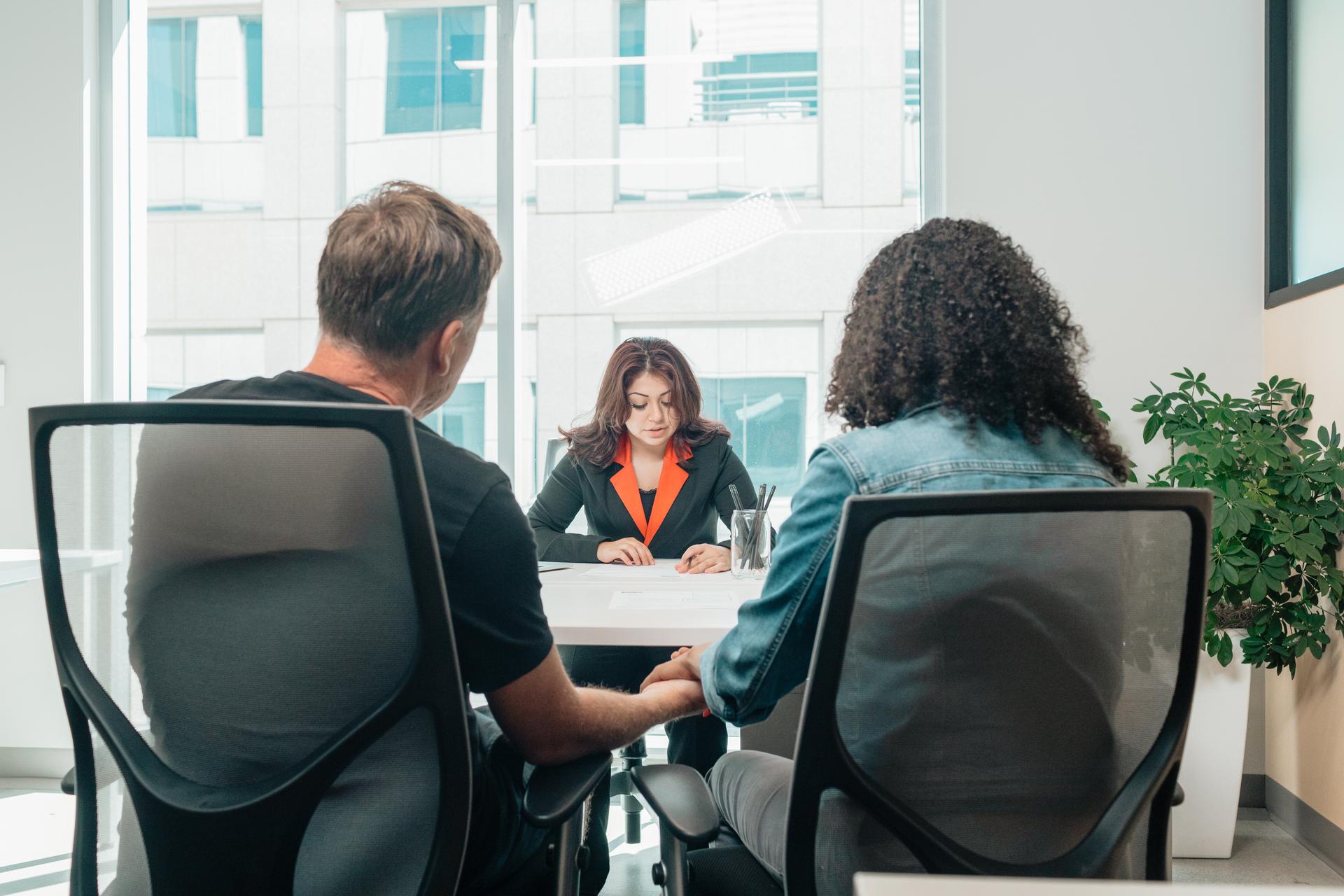 Two people seated at a table while a woman in a suit reads documents