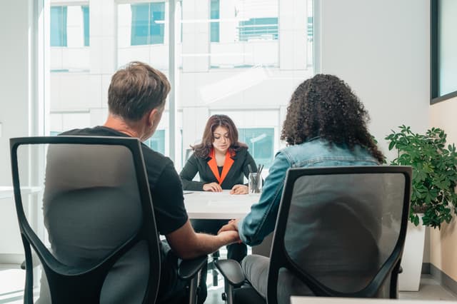 Two people seated at a table while a woman in a suit reads documents