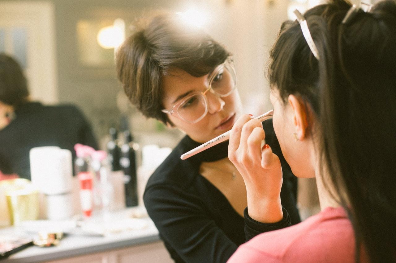 Makeup artist in glasses applying makeup with a brush to a client's face