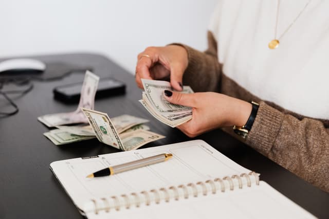 Hands counting US dollar bills beside an open planner, from the bookkeeper career guide