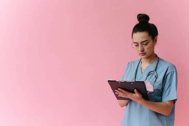 Nurse in blue scrubs reading a clipboard against a plain pink backdrop