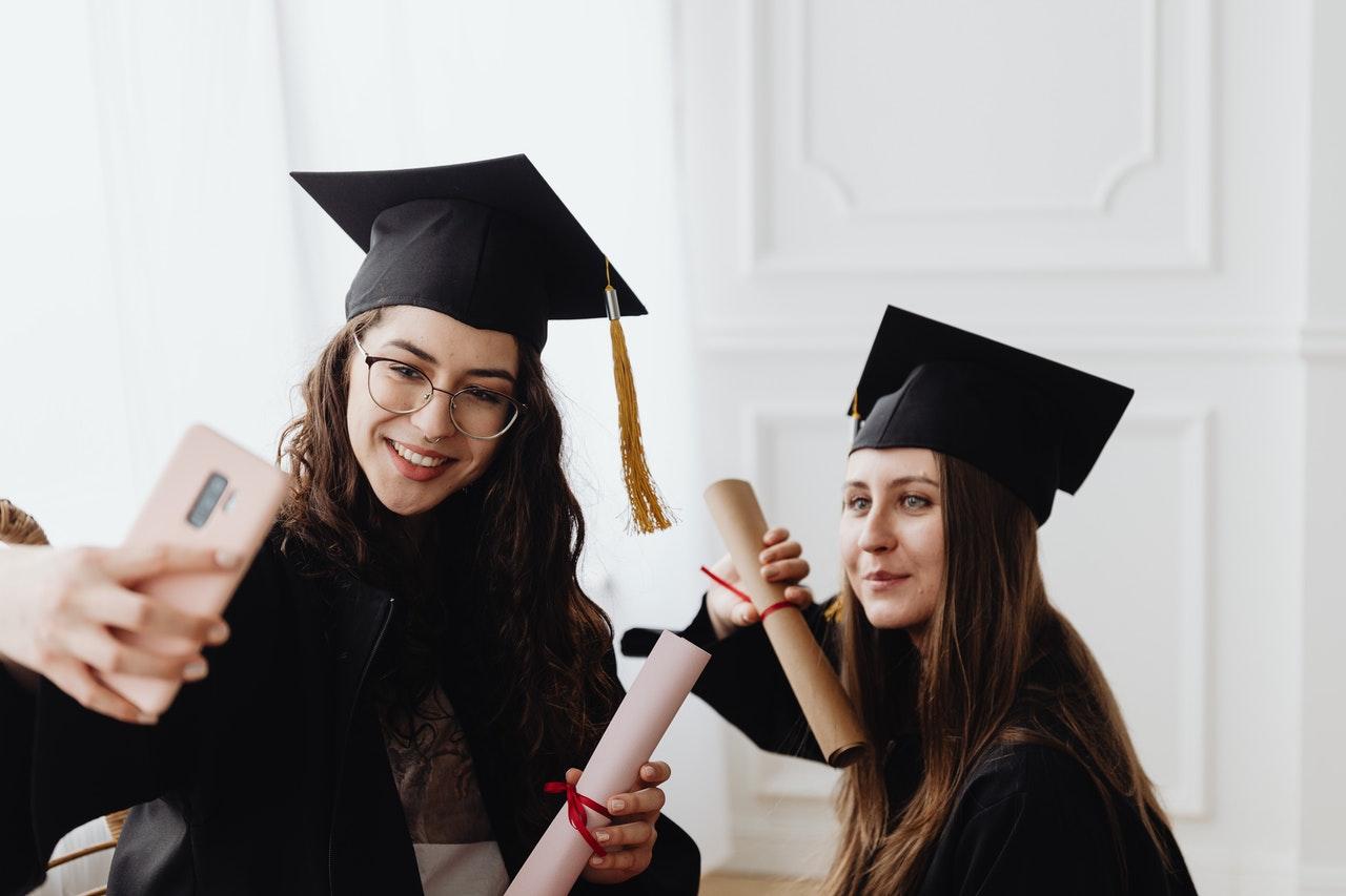 Two graduates in mortarboards take a selfie holding diplomas, from the college graduate salary guide