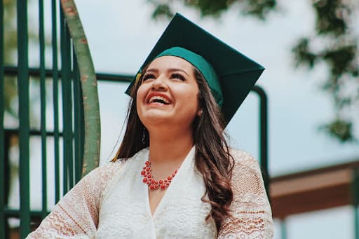 Laughing graduate in a green mortarboard and white lace top gazing upward outdoors