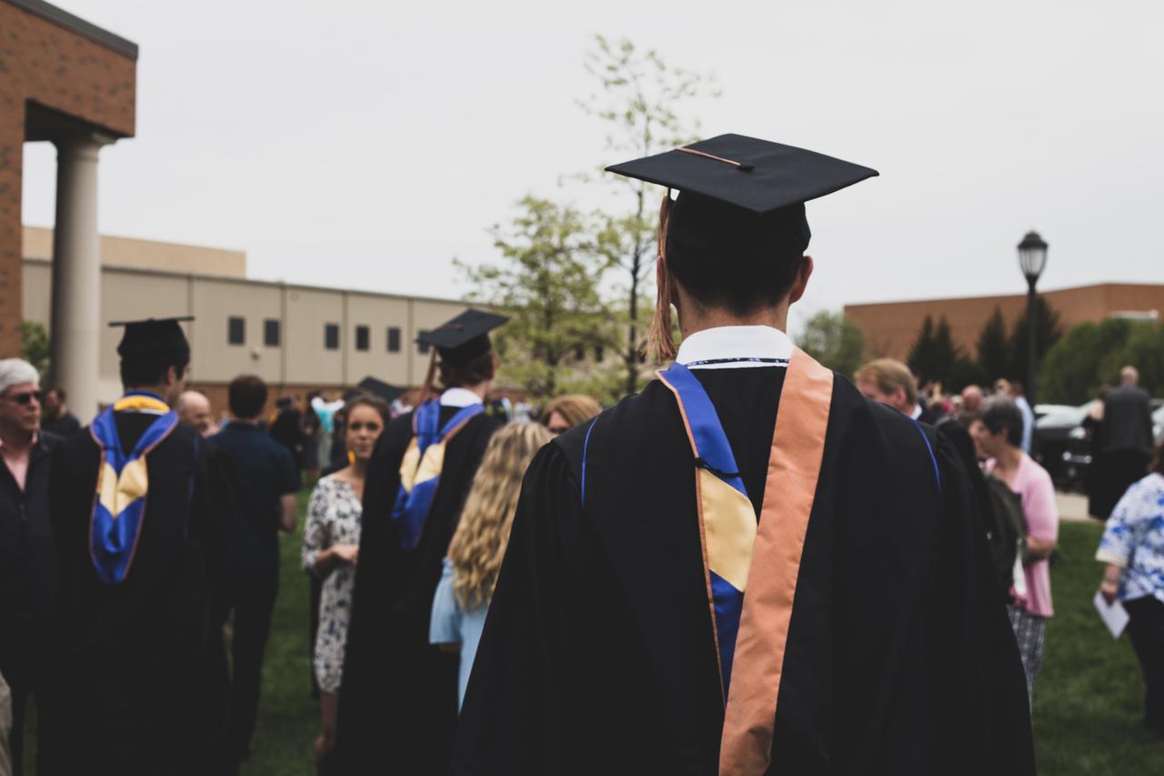 Graduate in mortarboard and hood seen from behind among a campus crowd