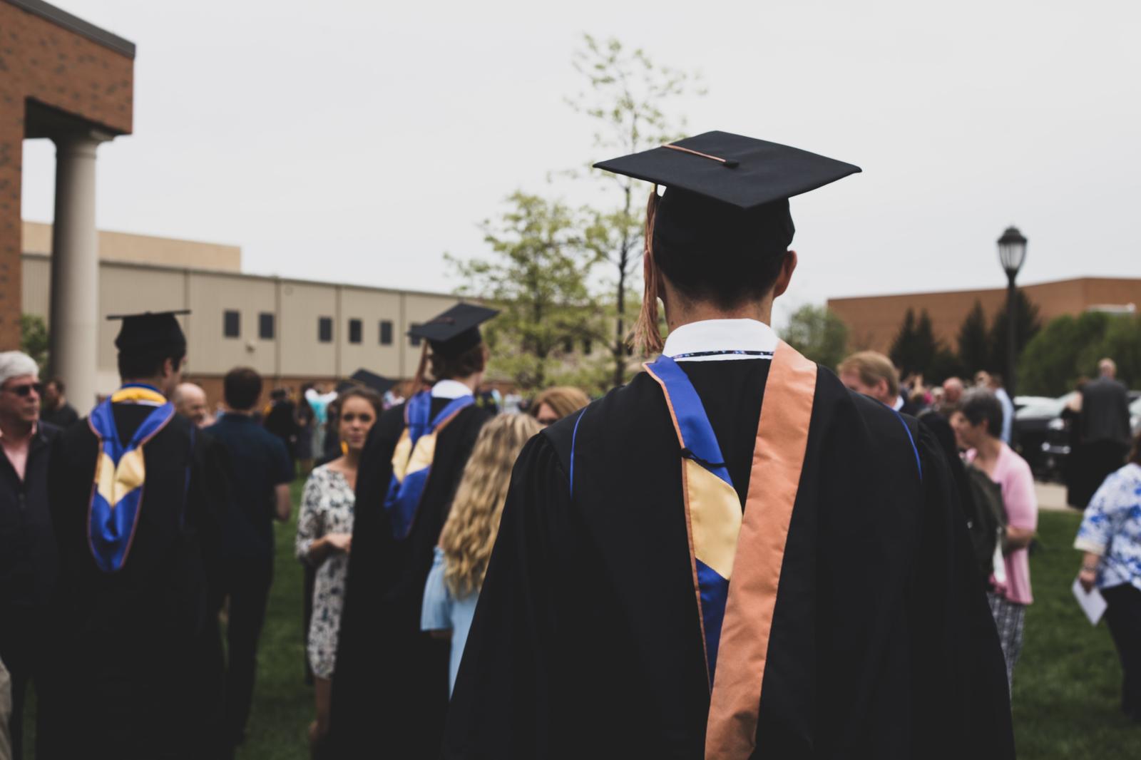 Graduate in mortarboard and hood seen from behind among a campus crowd