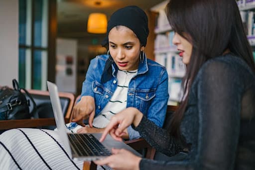 Two young women in a library pointing together at a laptop screen
