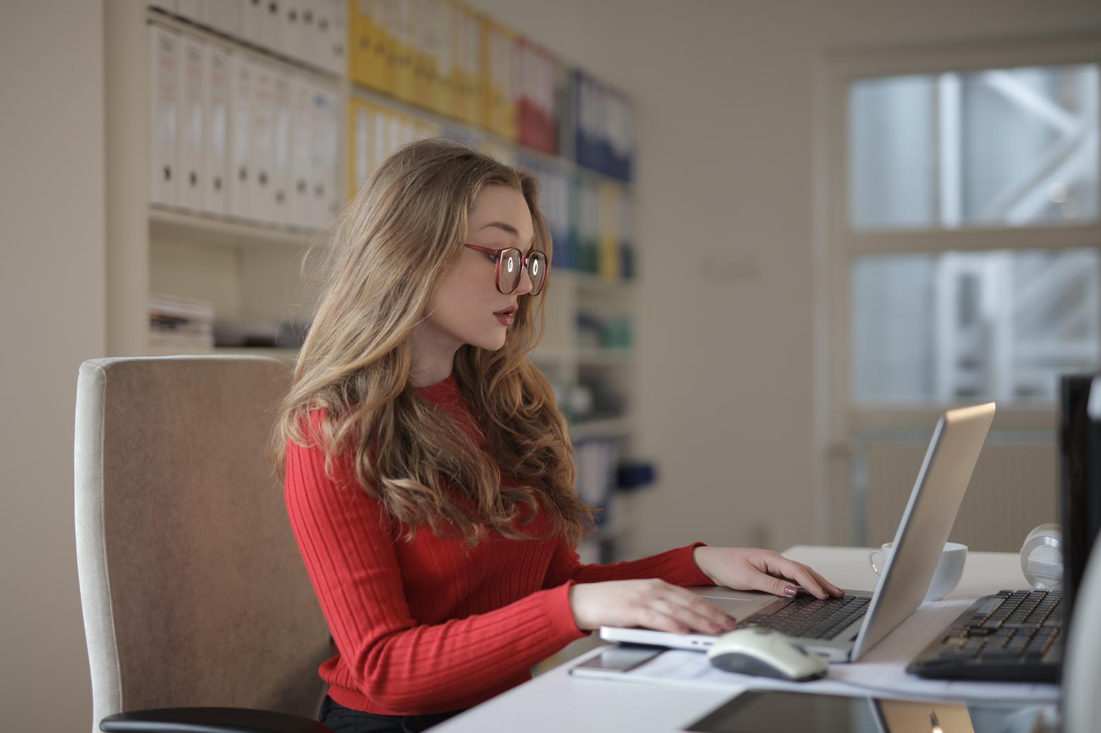 Woman in a red sweater typing on a laptop in a file-lined office