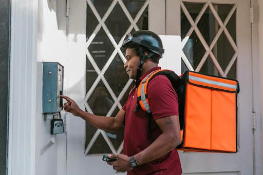 Delivery courier with an orange insulated backpack ringing a doorbell at a doorway