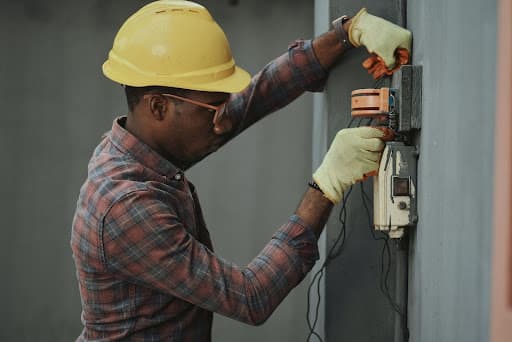 Man in a yellow hard hat and gloves wiring a wall-mounted electrical box