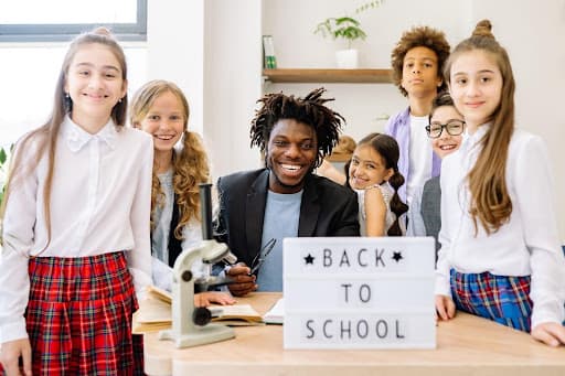 Grinning teacher and six schoolchildren behind a desk with a back to school sign