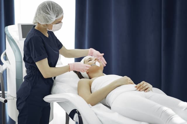 Esthetician in scrubs and gloves performing a facial treatment on a reclining client
