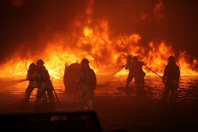 Firefighters silhouetted against a wall of flames, directing hose streams at the fire