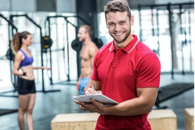 Smiling man in a red polo holds a clipboard and pen inside a gym