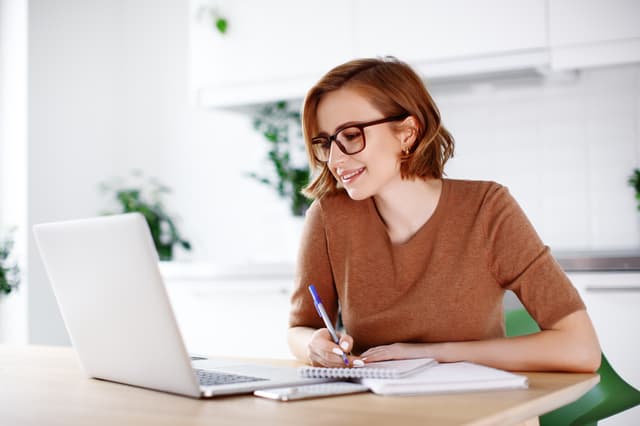 Woman in glasses writing notes in a notebook beside an open laptop