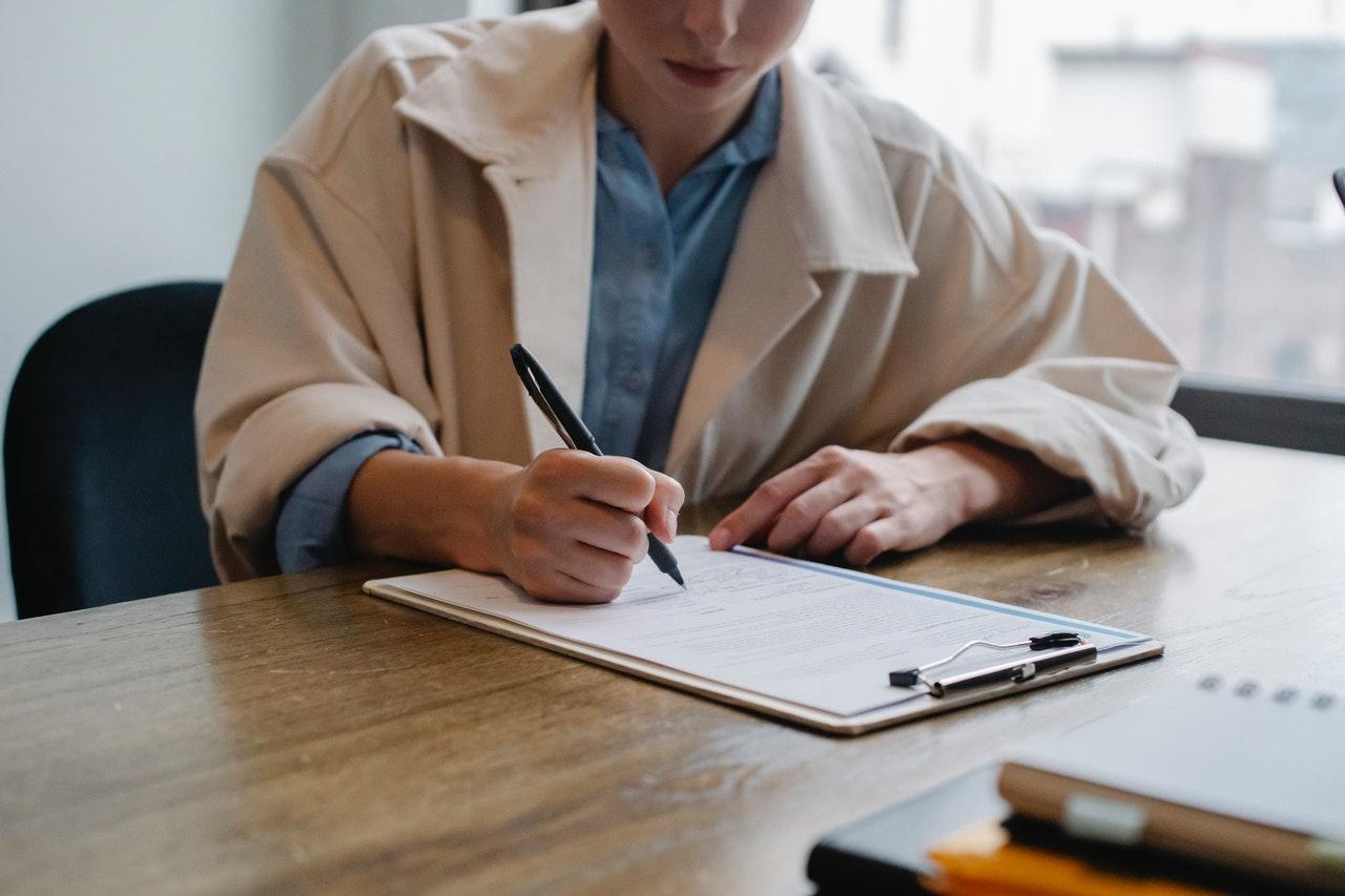 Woman in a beige blazer filling out a form on a clipboard at a desk