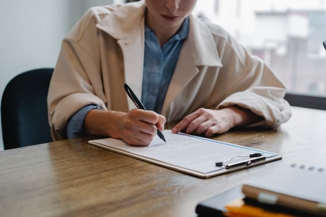 Woman in a beige blazer filling out a form on a clipboard at a desk