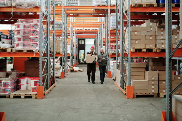 Two workers walking a warehouse aisle between stocked pallet racks, one carrying a box