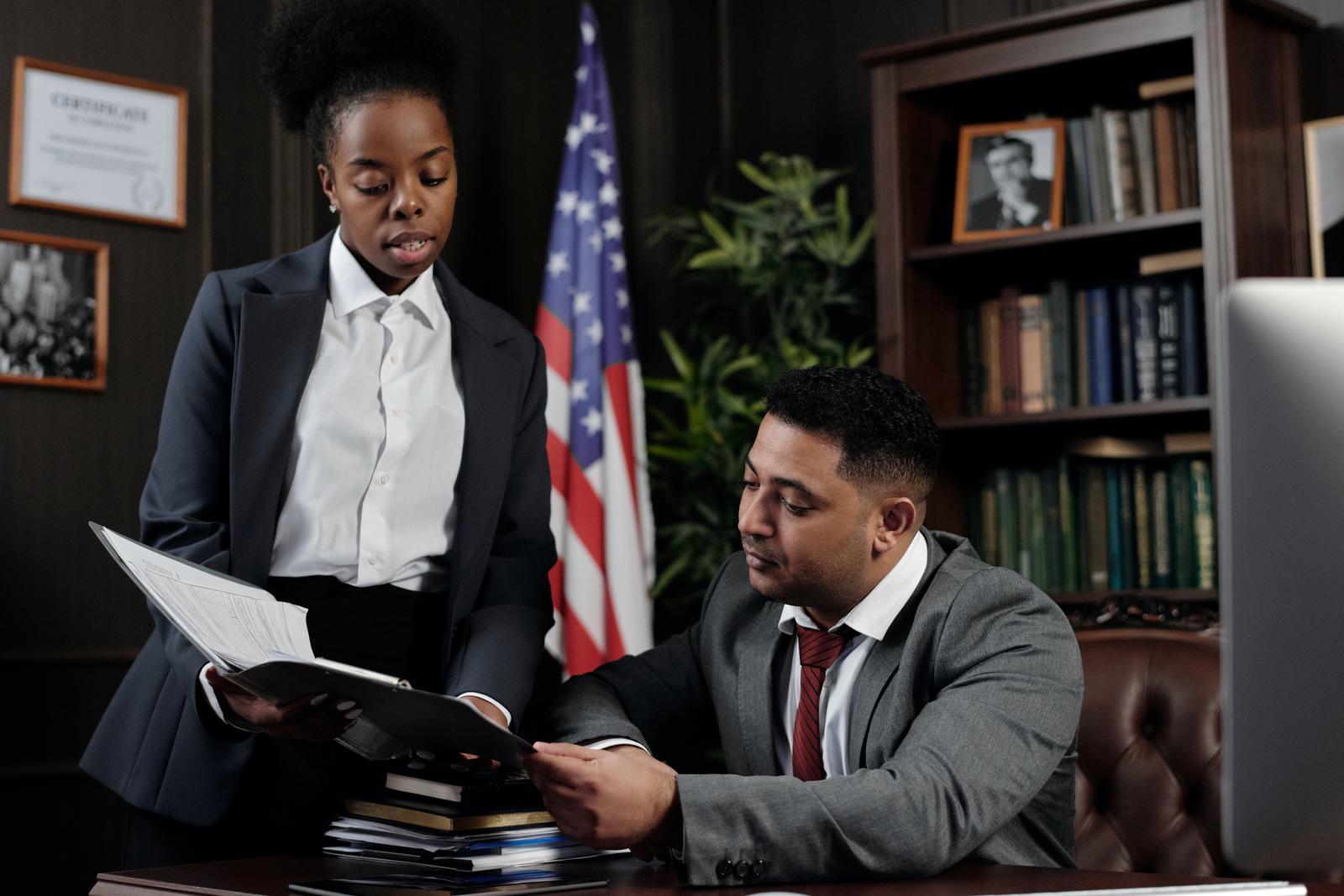 Two people in business suits review an open document folder beside a US flag