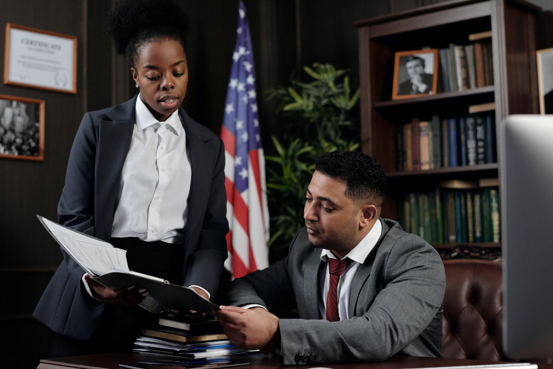 Two people in business suits review an open document folder beside a US flag
