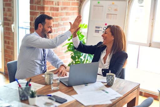 Colleagues in business attire high-fiving over a laptop and scattered paperwork