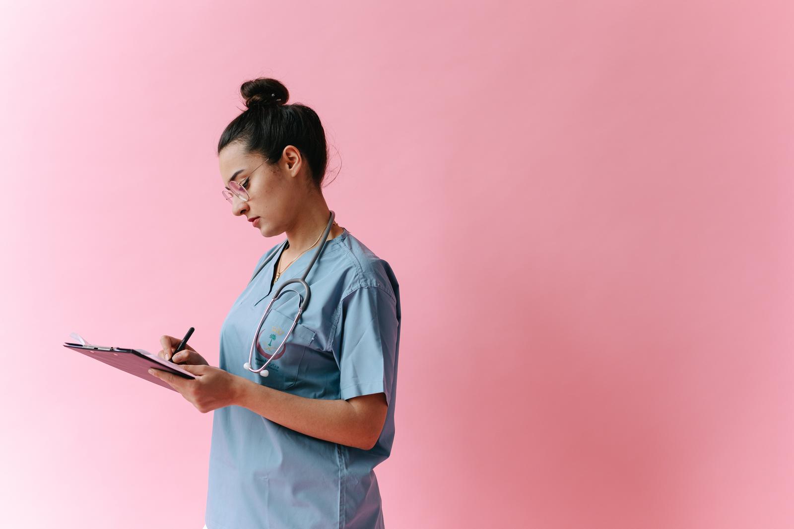 Healthcare worker in blue scrubs writing on a clipboard against a pink background
