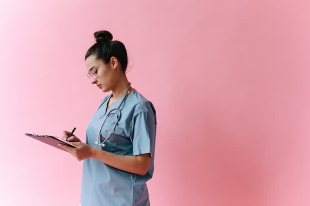 Healthcare worker in blue scrubs writing on a clipboard against a pink background