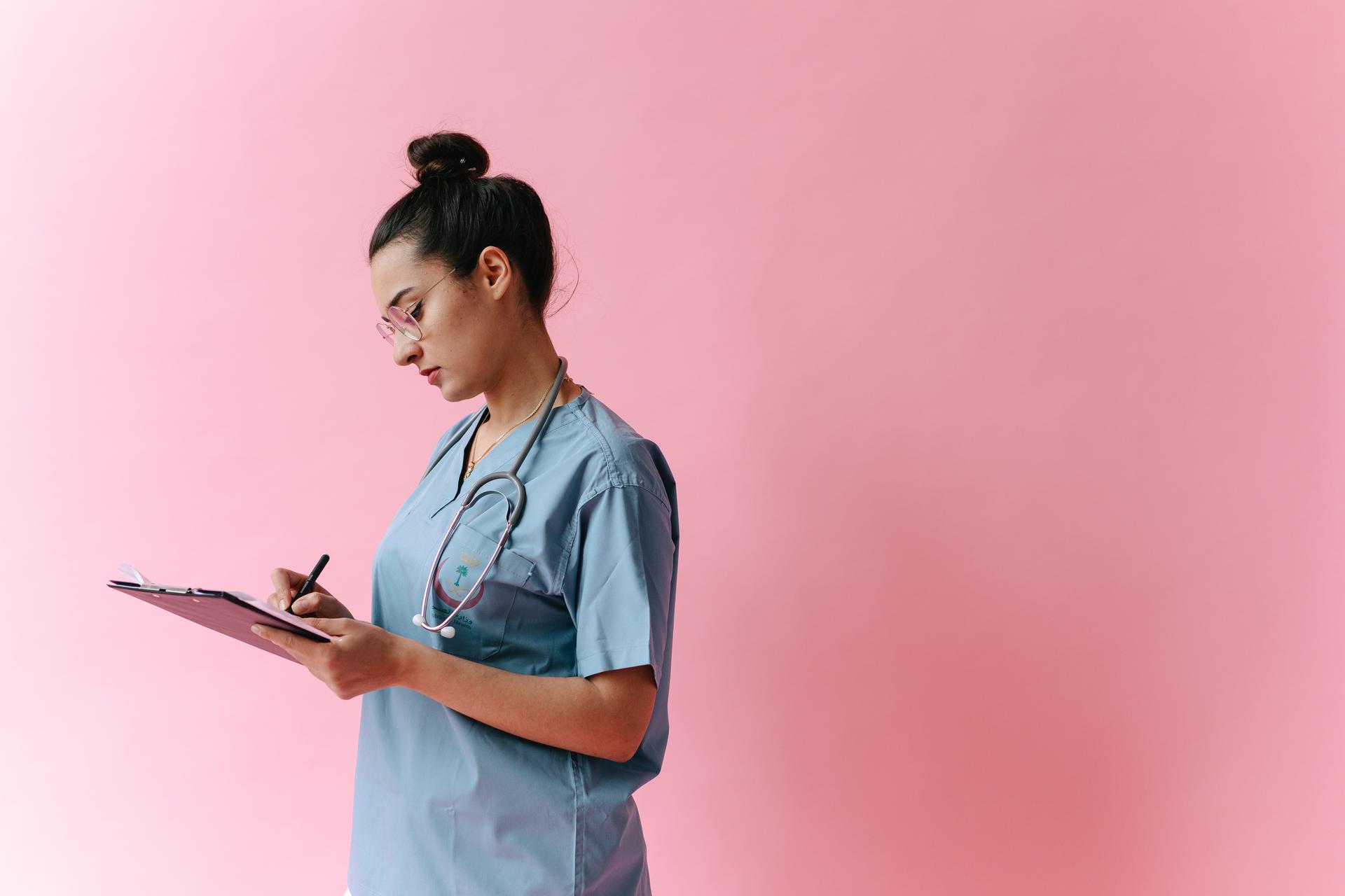 Healthcare worker in blue scrubs writing on a clipboard against a pink background