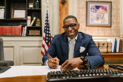 Smiling man in a suit signing a document at a desk beside an American flag