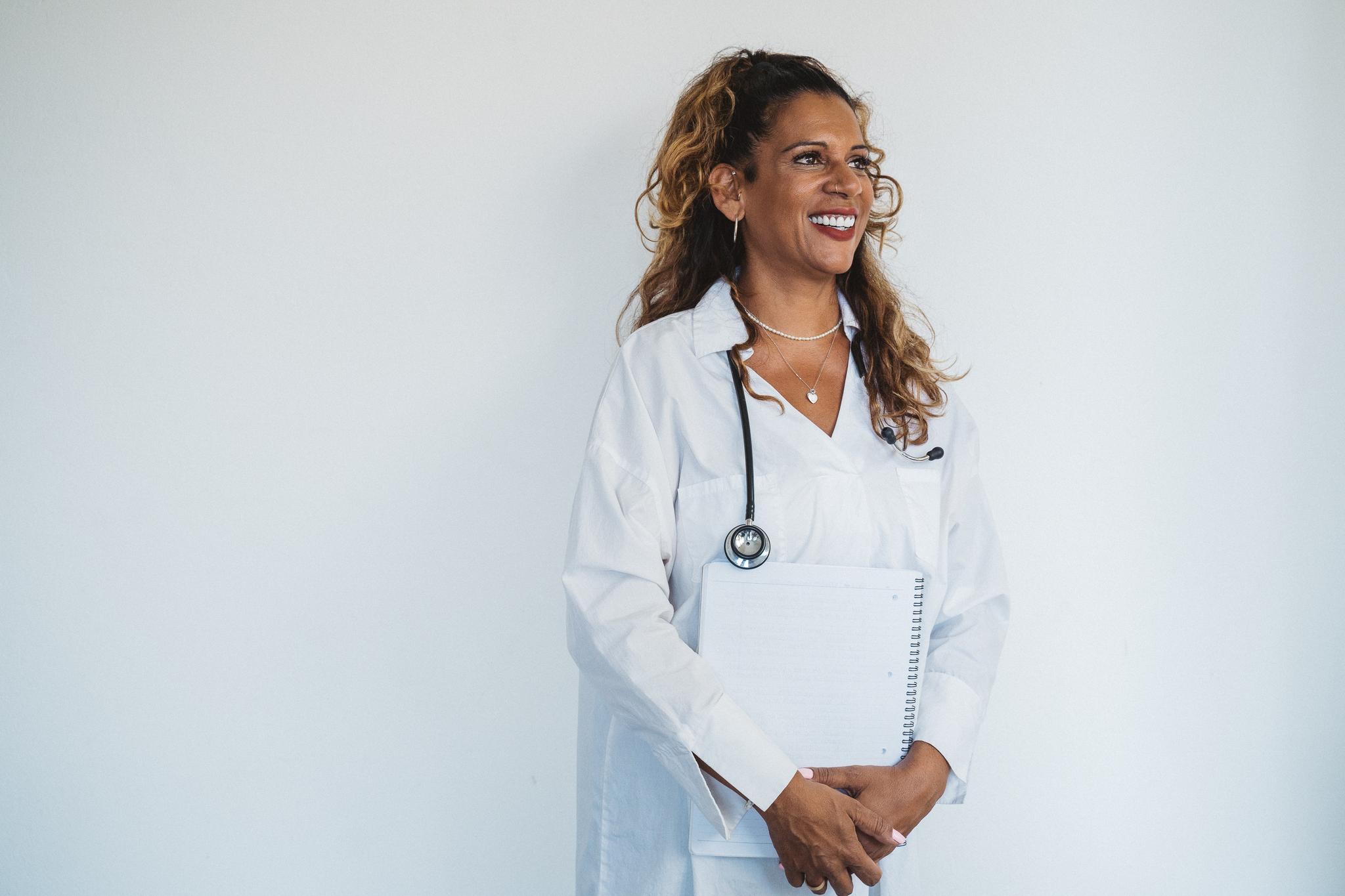 Smiling woman in a white coat with a stethoscope, holding a spiral notebook