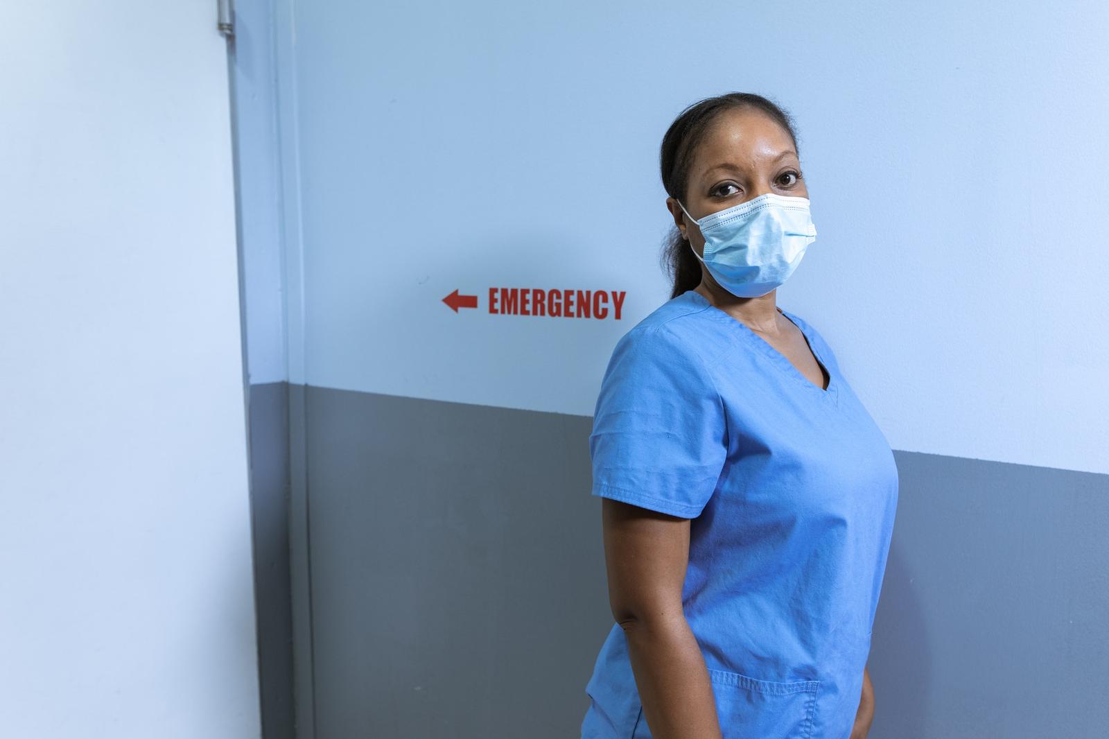 Nurse in blue scrubs and a face mask beside a red hospital emergency sign