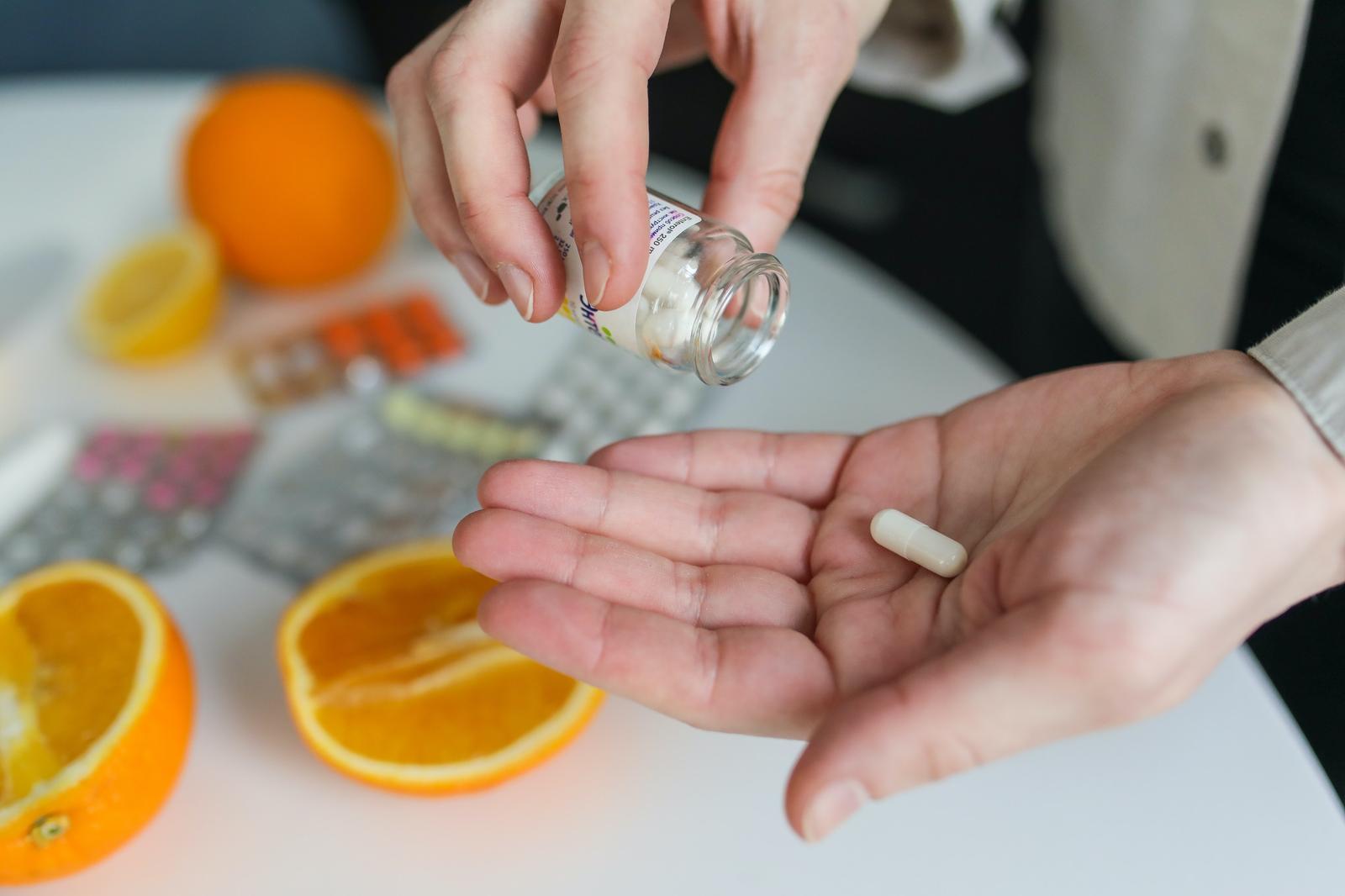 Hands tipping a capsule from a bottle into a palm beside blister packs and oranges