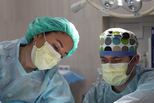 Two masked clinicians in surgical caps working beneath an operating room lamp