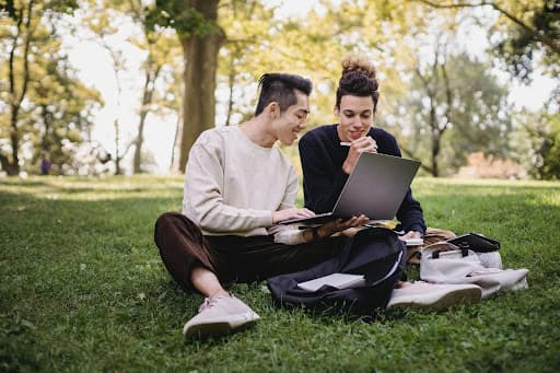 Two students sitting on park grass sharing a laptop with backpacks nearby