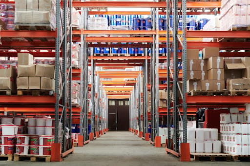 Long warehouse aisle of orange racking stacked with boxes, drums and pallets