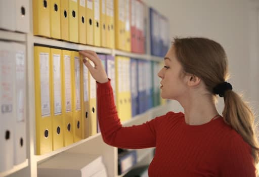 Woman in a red sweater pulling a yellow binder from crowded office shelves