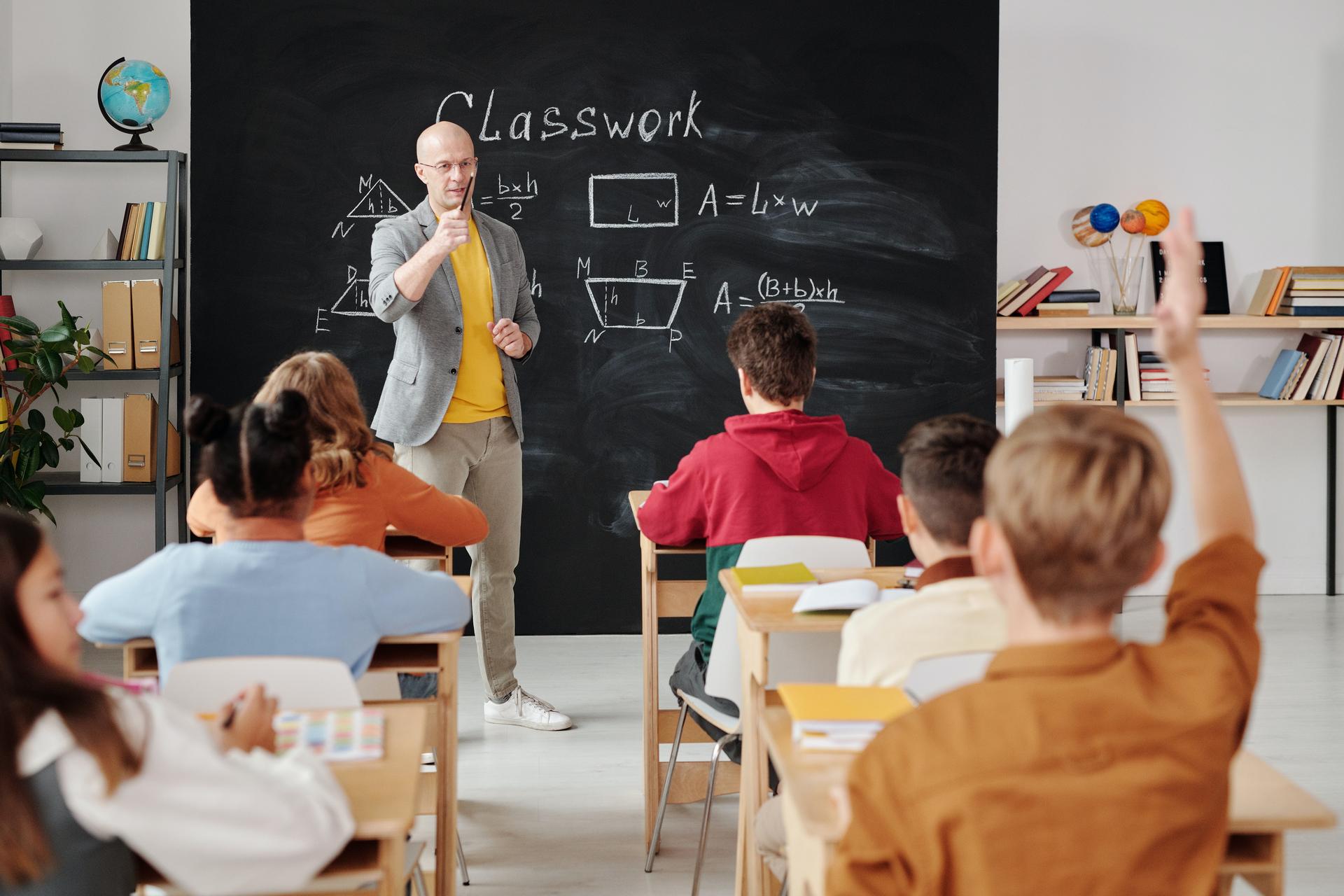 Teacher at a blackboard of area formulas as a seated student raises a hand