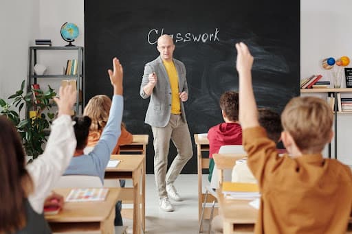 Teacher at a chalkboard marked Classwork as students raise their hands