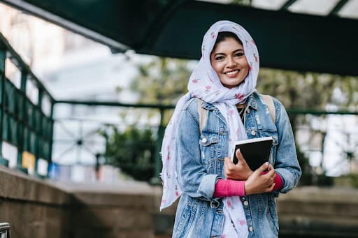 Student in a hijab and denim jacket holding a notebook outdoors, smiling