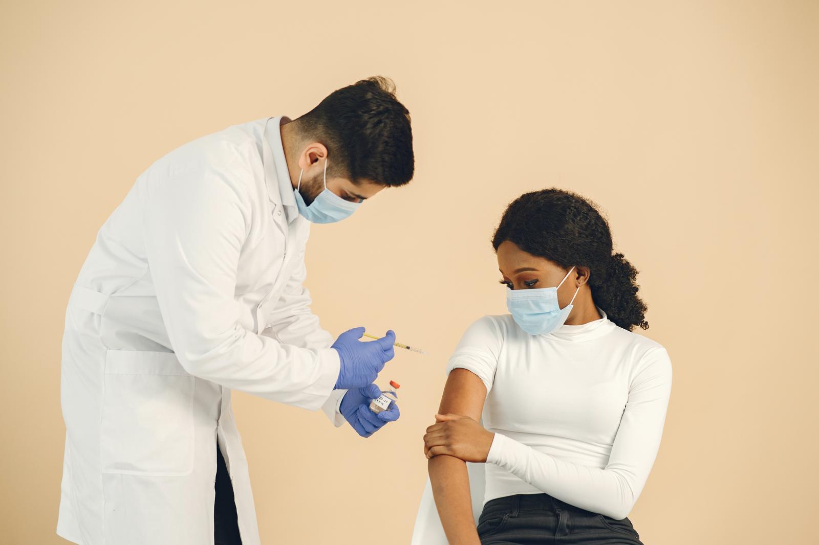 Masked clinician holding a syringe and vial beside a masked patient's bared upper arm