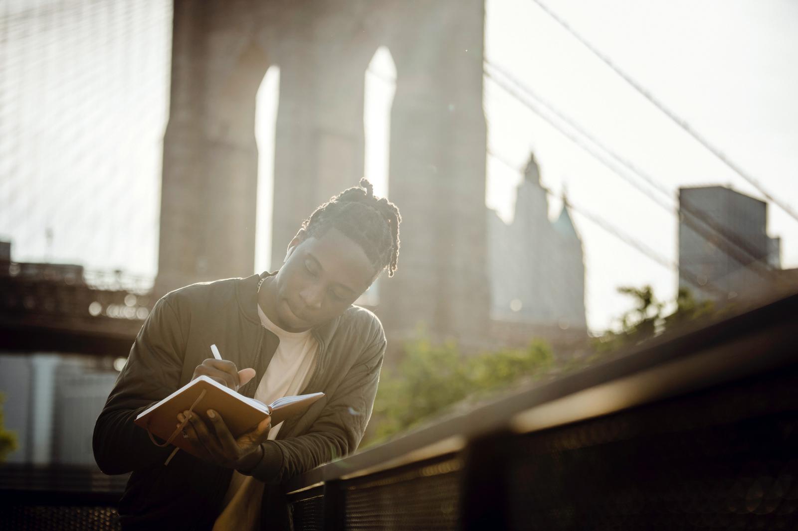 Thoughtful guy writing in notebook leaning on fence against bridge