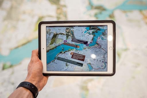 Hand holding a tablet showing an annotated Ottawa city map over paper maps