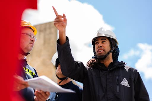 Workers in hard hats outdoors, one pointing skyward, another holding printed pages