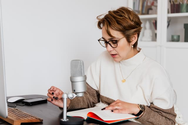 Voice actor reading aloud from a book into a studio microphone at a home desk