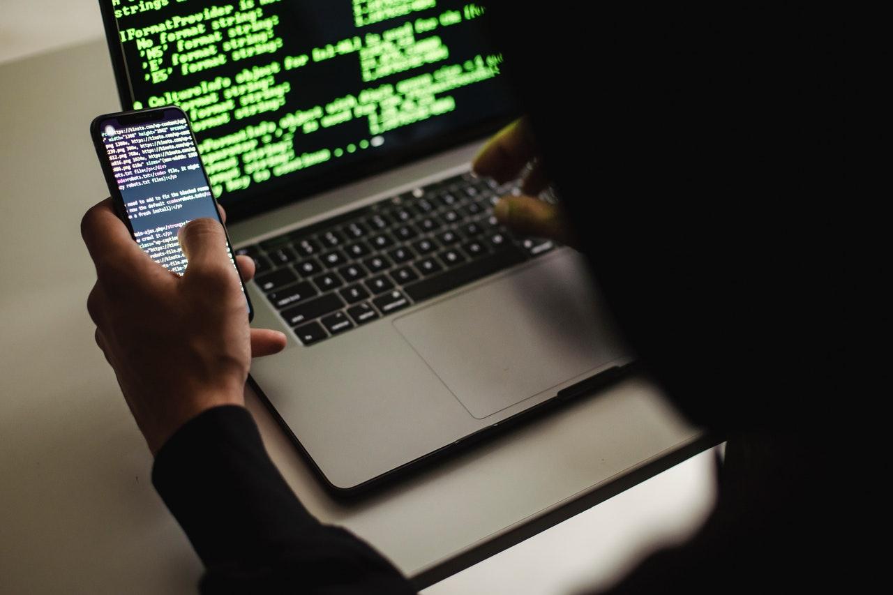 Developer holding a phone beside a laptop screen filled with green code
